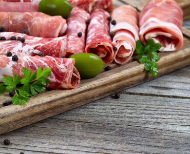 Close up horizontal image of various meats on serving board with ham pork beef parsley and olives on rustic wood. Focus on side part of serving board and first row of meat.