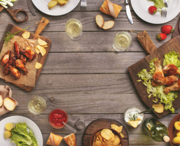 Outdoors Food Concept. On the wooden table different food with copy space grilled chicken legs buffalo wings bread salad potatoes strawberry and wine glasses with wine bottle